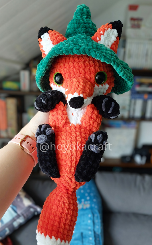Crocheted fox toy with a green hat held by a person in a casual indoor setting.